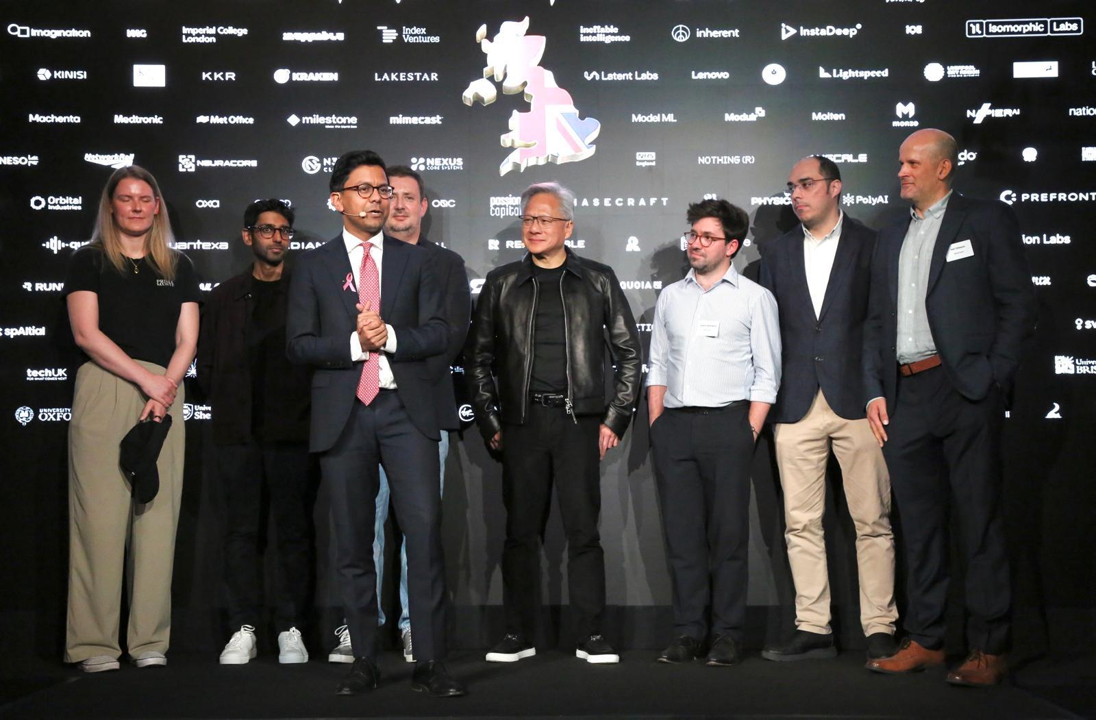 Britain's AI minister Kanishka Narayan, in a pink tie and headset microphone, speaks on stage beside Nvidia CEO Jensen Huang in his black leather jacket and a line of British tech founders, in front of a wall of UK company and university logos around a map of the UK